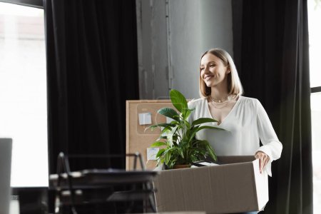 Happy new worker holding carton box with plant in office