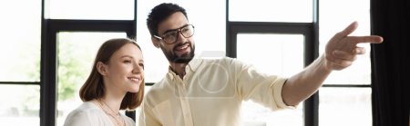 Cheerful businessman pointing with finger near intern in office, banner 