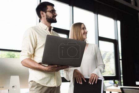 Téléchargez les photos : Homme d'affaires souriant avec ordinateur portable et stagiaire tenant dossier en papier dans le bureau - en image libre de droit