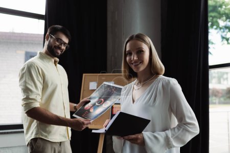 Smiling intern holding notebook and looking at camera near businessman with charts in office 