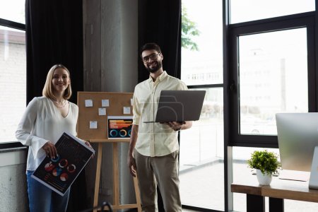Smiling manager holding laptop and looking at camera near intern with charts in office 