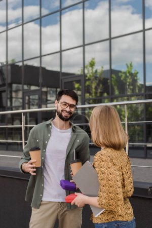 Hombre de negocios sonriente sosteniendo lonchera y café para llevar mientras habla con su colega con la carpeta de papel al aire libre 