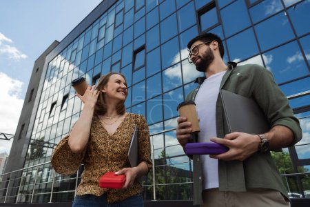 Low angle view of smiling businesswoman holding coffee to go and lunch box near colleague with laptop outdoors 