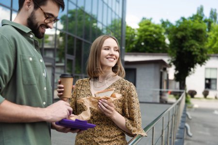 Femme d'affaires souriante tenant un sandwich et un café à emporter près d'un collègue dans une rue urbaine 