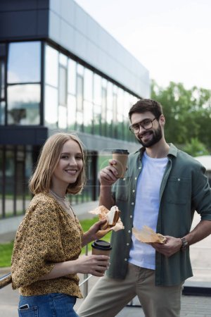 Businesswoman holding sandwich and coffee to go near colleague on urban street 
