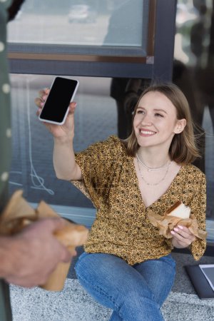 Mujer de negocios sonriente sosteniendo sándwich y teléfono inteligente cerca de colega en la calle urbana 