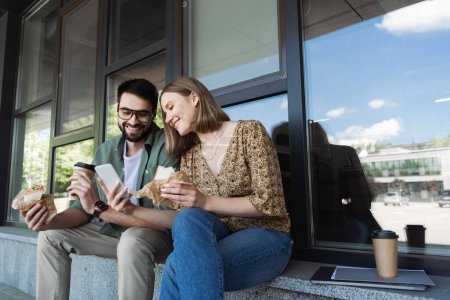 Mujer de negocios sonriente mostrando smartphone a colega con sándwich y café para salir al aire libre 