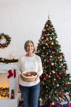 heureuse femme d'âge moyen avec les cheveux gris tenant panier en osier et des boules près de l'arbre de Noël 