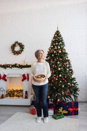 glückliche Frau mittleren Alters mit grauen Haaren, die Weidenkorb und Christbaumkugeln in der Nähe des Weihnachtsbaums mit Geschenken hält 