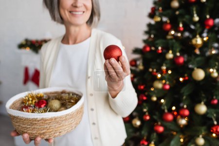 cropped view of cheerful middle aged woman with grey hair holding wicker basket and bauble near christmas tree 