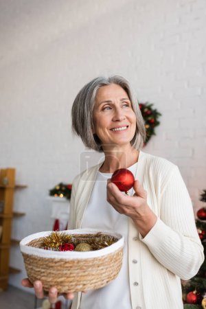 glückliche Frau mittleren Alters mit grauen Haaren hält Weidenkorb und rote Weihnachtskugel 