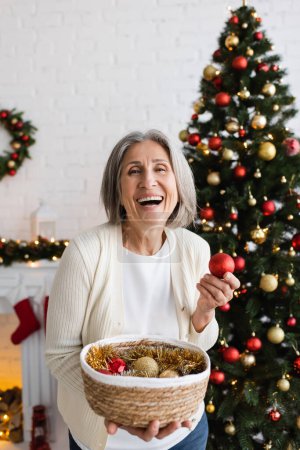 fröhliche Frau mittleren Alters mit grauen Haaren, die Weidenkorb und Christbaumkugeln in der Nähe des Weihnachtsbaums hält 