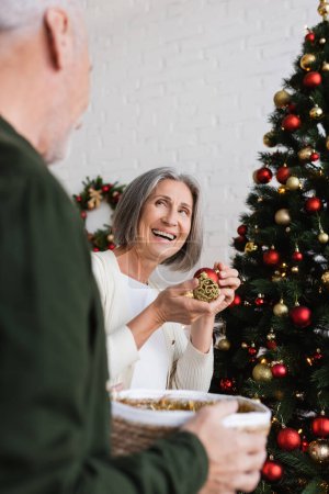Téléchargez les photos : Joyeuse femme d'âge moyen avec des cheveux gris décorant arbre de Noël près du mari - en image libre de droit