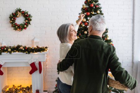 smiling middle aged woman decorating christmas tree and looking at husband holding wicker basket 