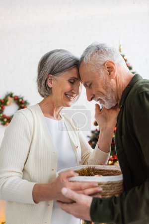 glückliches älteres Paar mit geschlossenen Augen hält Weidenkorb in der Nähe geschmückten Weihnachtsbaum