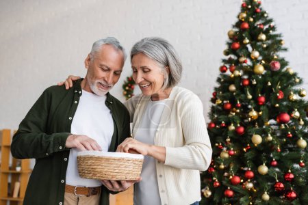 smiling middle aged couple looking at wicker basket near decorated christmas tree
