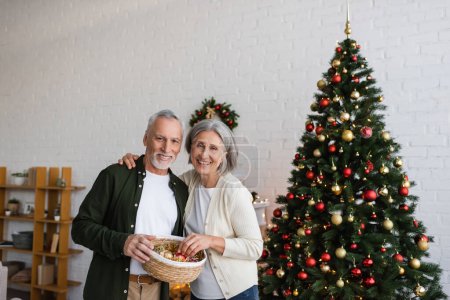 smiling middle aged couple holding wicker basket with baubles near decorated christmas tree