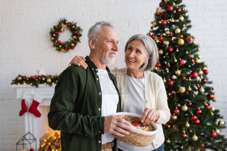smiling middle aged couple holding wicker basket with baubles near decorated pine tree