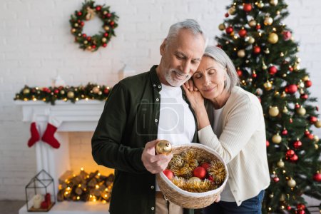 fröhliche Frau mittleren Alters lehnt an Mann mit Weidenkorb und schaut Weihnachtskugel an