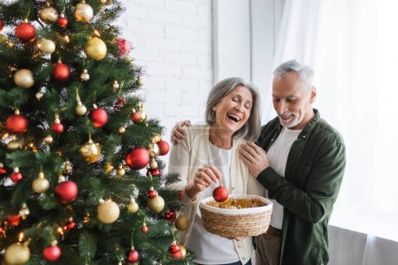 cheerful middle aged woman laughing while holding bauble near husband and christmas tree