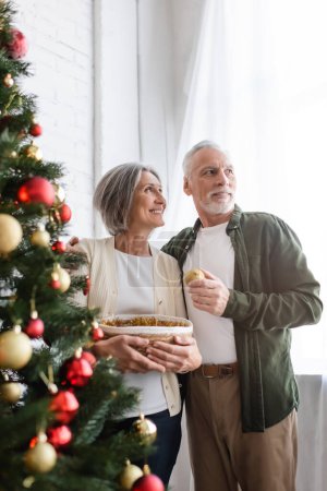 Téléchargez les photos : Joyeux mari d'âge moyen étreignant femme et regardant loin près décoré arbre de Noël - en image libre de droit