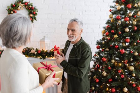 smiling middle aged man holding present and looking at wife near christmas tree