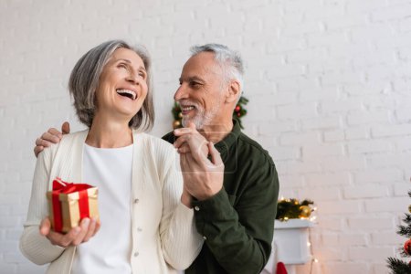 cheerful mature man holding hand of happy wife with christmas present