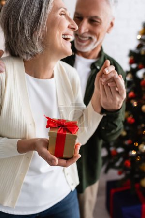 cheerful mature man holding hand of joyful wife with christmas present laughing at home