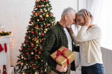 bearded middle aged man holding present and hugging with smiling wife near christmas tree