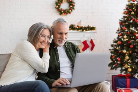Téléchargez les photos : Heureux couple d'âge moyen regarder film sur ordinateur portable près décoré arbre de Noël - en image libre de droit
