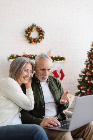 middle aged man pointing at laptop near wife in glasses and christmas tree 