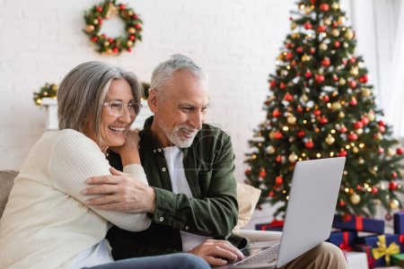 Téléchargez les photos : Sourire couple d'âge moyen regarder film comédie sur ordinateur portable près décoré arbre de Noël - en image libre de droit