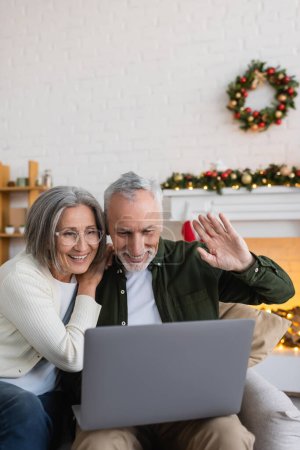 Téléchargez les photos : Souriant homme d'âge moyen agitant la main près de la femme tout en ayant appel vidéo sur ordinateur portable pendant Noël - en image libre de droit