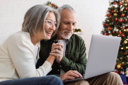 Téléchargez les photos : Sourire couple d'âge moyen tenant la main et ayant appel vidéo sur ordinateur portable pendant Noël - en image libre de droit