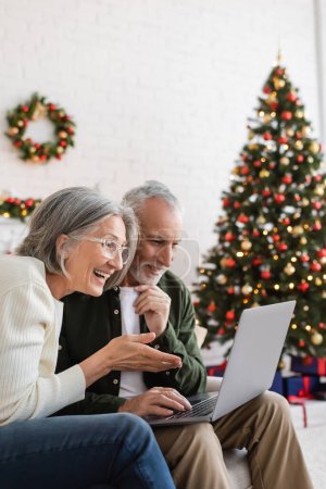 Téléchargez les photos : Souriant femme d'âge moyen geste près du mari et avoir un appel vidéo sur ordinateur portable pendant Noël - en image libre de droit