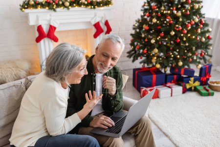 Téléchargez les photos : Souriant femme d'âge moyen pointant vers mari heureux et ayant appel vidéo sur ordinateur portable pendant Noël - en image libre de droit