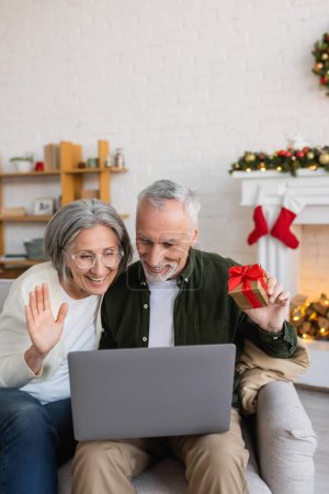sonriente mujer de mediana edad saludando mano cerca de marido y tener videollamada en el ordenador portátil durante la Navidad 