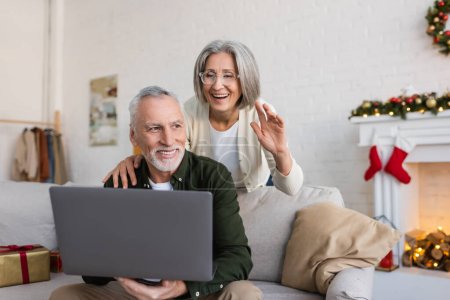 cheerful middle aged woman in glasses waving hand near husband during video call on christmas day
