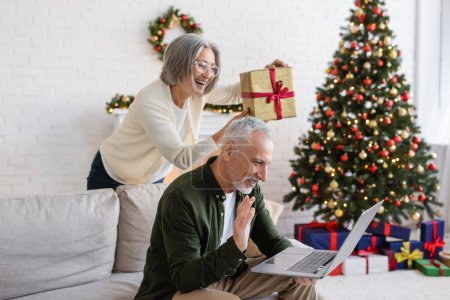 fröhliche Frau mittleren Alters in Brille zeigt Weihnachtsgeschenk in der Nähe ihres Mannes während eines Videogesprächs 