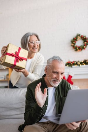 cheerful middle aged woman in glasses showing christmas present near husband waving hand during video call 