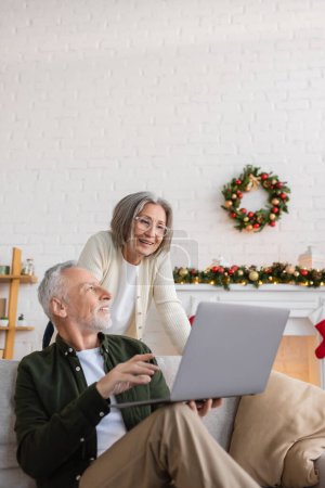 cheerful middle aged man showing laptop to wife in glasses on christmas day 