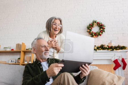 fröhlicher Mann mittleren Alters zeigt seiner geschmeichelten Frau an Heiligabend Laptop in Brille 
