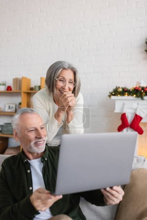 Téléchargez les photos : Heureux homme d'âge moyen montrant ordinateur portable à flatté femme dans des lunettes le jour de Noël - en image libre de droit