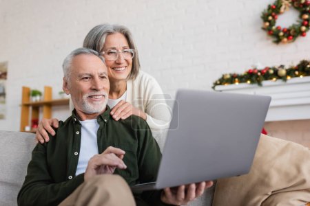 heureuse femme d'âge moyen dans des lunettes étreignant mari et regardant ordinateur portable pendant appel vidéo le jour de Noël 