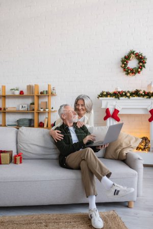 mujer de mediana edad feliz en gafas abrazando marido sentado en el sofá con el ordenador portátil en el día de Navidad 