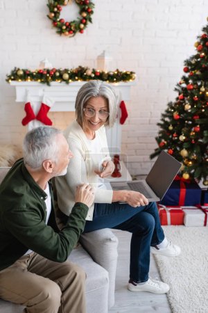 Téléchargez les photos : Femme d'âge moyen dans des lunettes en utilisant un ordinateur portable près mari barbu pendant les vacances de Noël - en image libre de droit