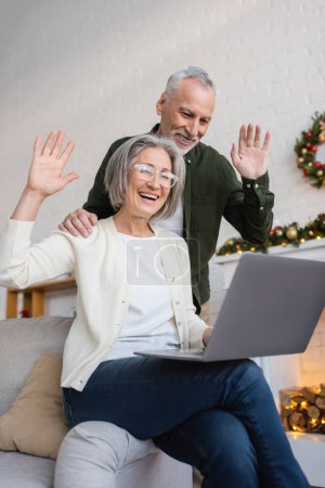 Téléchargez les photos : Heureux mari et femme d'âge moyen agitant les mains pendant l'appel vidéo pendant les vacances de Noël - en image libre de droit