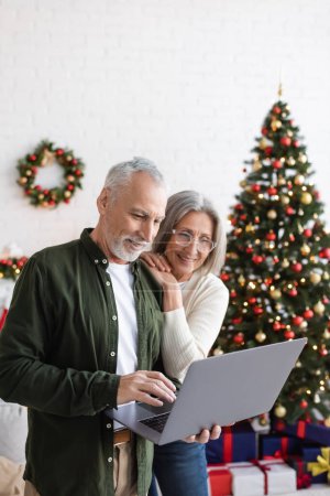 Téléchargez les photos : Joyeux mari et femme d'âge moyen regardant ordinateur portable près décoré arbre de Noël - en image libre de droit