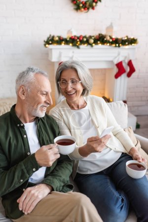 cheerful and mature woman in glasses holding smartphone near husband with cup of tea during christmas holidays 