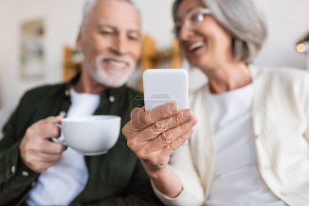 Téléchargez les photos : Femme gaie et mature tenant smartphone près mari heureux avec tasse de thé - en image libre de droit